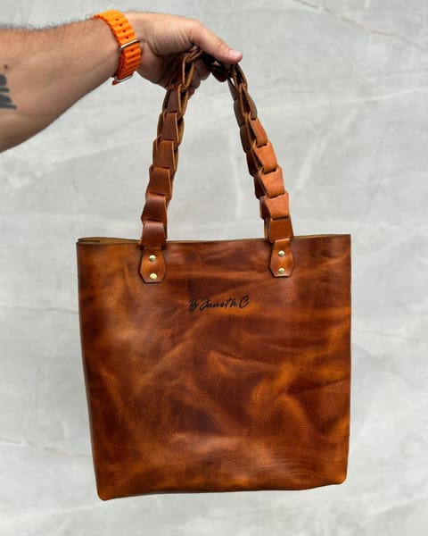 Bolso tote de cuero miel en la mano, con las asas trenzadas y un nombre grabado