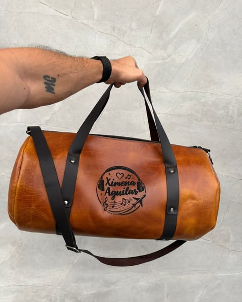 Maleta de cuero miel en la mano, con correas café y un nombre con un diseño de música grabados