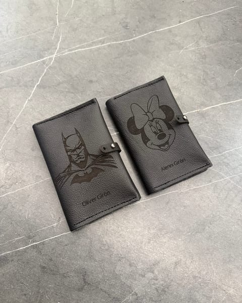 Dos portapasaportes dobles de cuero negro, con personajes de dibujos animados y nombres grabados