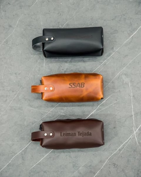 Tres travel dopp de cuero negro, miel y café, con nombres y un logo grabados