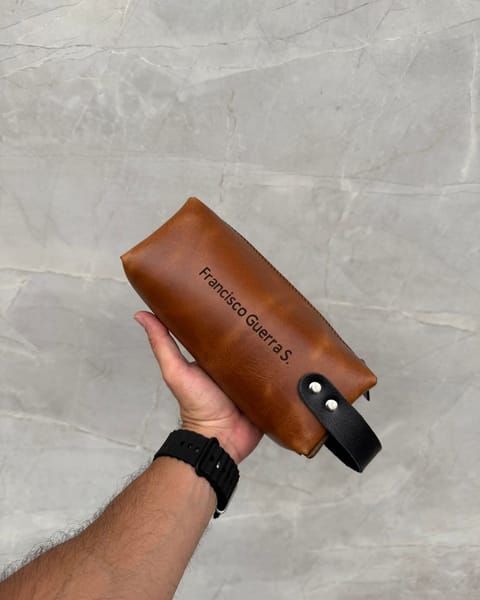 Travel dopp de cuero miel en la mano, con correa negra y un nombre grabado